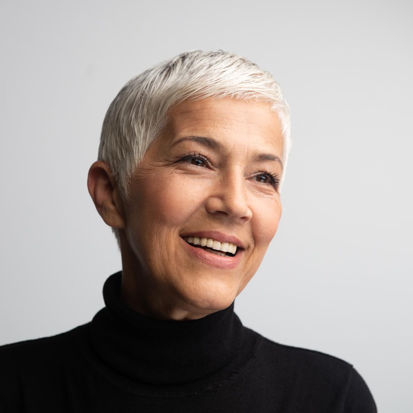 Smiling older woman with short silver hair wearing a black turtleneck, representing natural rejuvenation and the artistry of facelift surgery by Dr. Monica Tadros in Englewood.